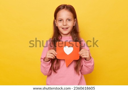 Popular child content, blogging. Portrait of charming preschool girl holding heart like icon of social media and looking at camera with toothy smile isolated over yellow background