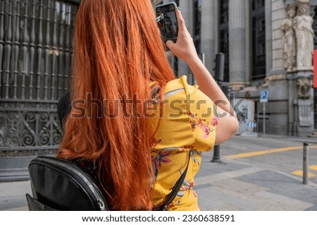 Red-haired girl with straight hair and a yellow dress taking a photo with her mobile phone in the streets of Madrid. Royalty-Free Stock Photo #2360638591