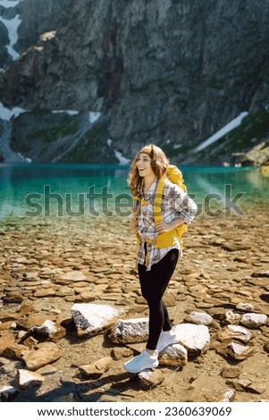 A smiling female traveler with a yellow hiking backpack against the backdrop of a turquoise lake among the mountains. Active lifestyle. Hiking, adventure concept.