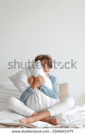 Good sleeping, time to sleep, youth daily health habit concept. Teenager boy sitting on bed, embracing pillow and smiling. Empty white wall background. Royalty-Free Stock Photo #2360641479