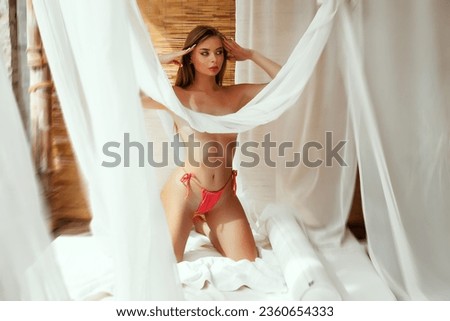 Pretty woman in pink knitted bikini without bra, cover breast white curtains enjoying resort vacation on bed with transparent curtains.