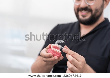 Cheerful orthodontist putting transparent aligners on artificial lower jaw in modern stomatology clinic bearded dentist presenting device for teeth straightening Royalty-Free Stock Photo #2360672419