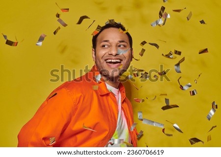 cheerful indian man in bright orange jacket smiling near falling confetti on yellow backdrop