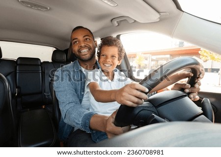 Fatherhood, parenthood, family, affection. Happy african american young father and preteen boy son driving car together. Child sit on dad laps, hold hands on steering wheel, bonding with his daddy