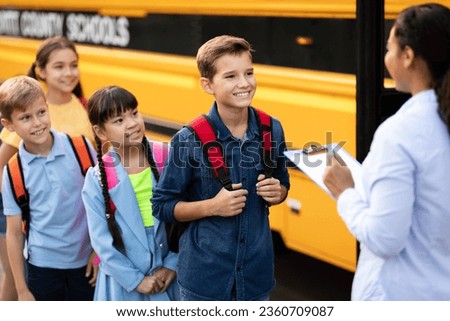 Happy schoolchildren waiting to get on school bus, standing in turn, friendly african american teacher holding clipboard, updating check list of children entering vehicle, selective focus Royalty-Free Stock Photo #2360709087