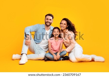 Parenthood and childhood concept. Happy caucasian parents mom and dad sitting on floor with daughter, posing isolated on yellow background and smiling at camera