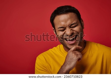 joyous indian man in yellow t-shirt smiling with closed eyes on red backdrop in studio, portrait Royalty-Free Stock Photo #2360710823