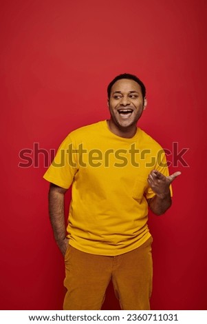 smiling indian man in bright casual clothes mocking and pointing with finger on red background Royalty-Free Stock Photo #2360711031