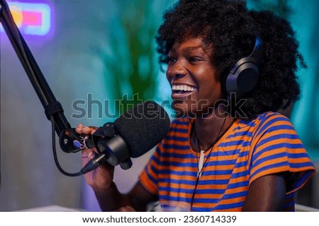 A portrait of a cheerful interracial podcaster is sitting in a broadcasting studio at home with headphones on and speaking at the microphone. A happy journalist is discussing live on air on a podcast.