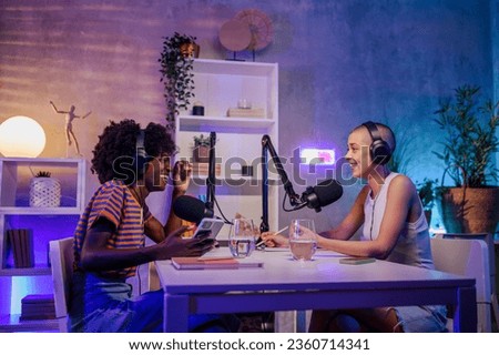 Two interracial podcasters are sitting in a home broadcasting studio and filming a podcast while going live on air and streaming. Two interracial gen z girls discussing funny topics on a podcast.