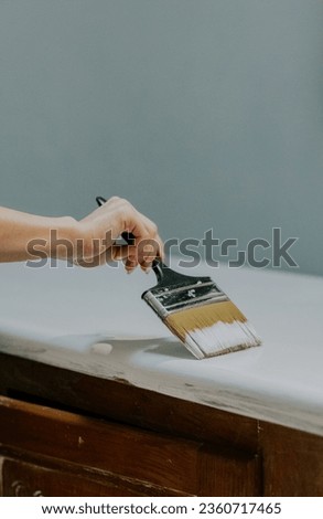 Young Caucasian unrecognizable girl paints an old cabinet with one hand in her room with a wide brush and white paint, close-up side view.