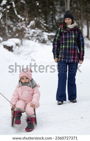 Vertical image showing a little girl sitting on a wood sled looking at the camera with her aunt behind her and smiling at her walking in the middle of the snowy winter mountains. Christmas holidays.