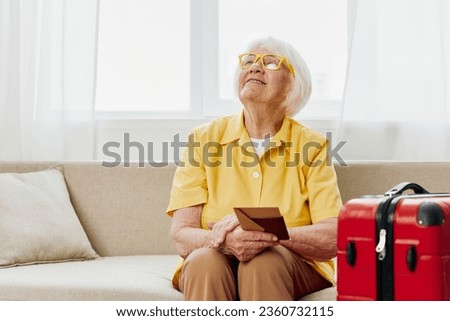 Happy senior woman with passport and travel ticket packed a red suitcase, vacation and health care. Smiling old woman joyfully sitting on the sofa before the trip raised her hands up in joy. Royalty-Free Stock Photo #2360732115