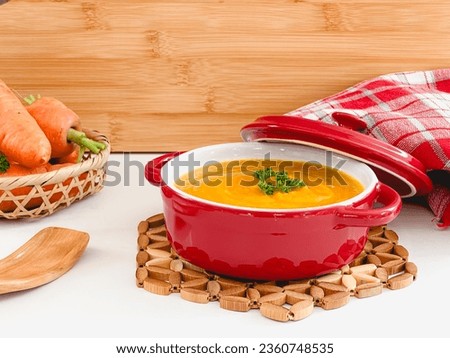 Cream of carrot soup, in red pot, on a white countertop and wooden background, copy space Royalty-Free Stock Photo #2360748535