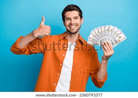 Photo of excited confident man dressed orange shirt rising cash fan showing thumb up isolated blue color background