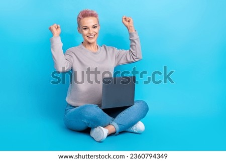 Full size photo of delighted cheerful girl sit floor use netbook raise fists success empty space isolated on blue color background
