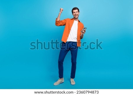 Full size photo of excited delighted guy hold smart phone raise fist achievement shout yes hooray isolated on blue color background