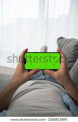 Young man at Home Uses Green Mockup Screen Smartphone in His Cozy Living Room.Vertical shot.Streaming service,watch videos online