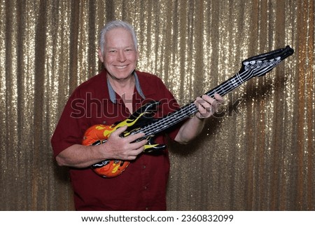 Photo Booth. A man smiles and poses like a Rock Star with an Inflatable Guitar while waiting to have his pictures taken in a Photo Booth at a Wedding Party. Photo Booth Props. Guitar. Rock Star. FUN. 