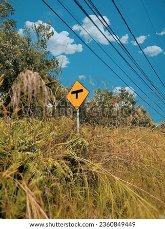 T-junction traffic sign stands among wild grass and trees under a clear sky and stretched power lines