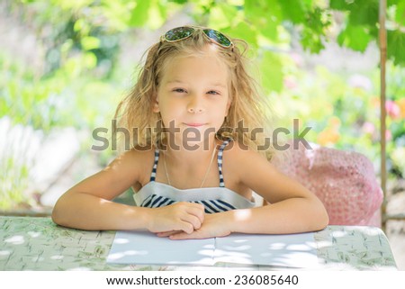 Little girl talking Silit at table and wrote against green of Park in summer.