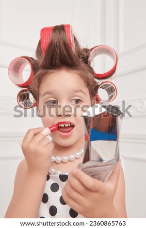 Kid's Fashion - clothes and accessories. A little pretty girl in an elegant polka-dot dress and curlers on her head is going to a party and paints her lips with lipstick. Pin-up style.