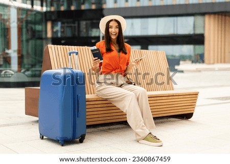 Excited Woman Passenger Utilizing Mobile App On Phone to Text, Browse and Book Tickets Online In Travel Application, Comfortably Seated with Suitcase Outdoors. Lady Using Cellphone Waiting Flight Royalty-Free Stock Photo #2360867459