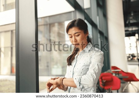 Businesswoman looking on her wrist watch while standing near office window. High quality photo Royalty-Free Stock Photo #2360870413