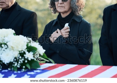 Cropped shot of mature woman with handkerchief and other people in mourning attire standing by coffin covered with USA flag at funeral