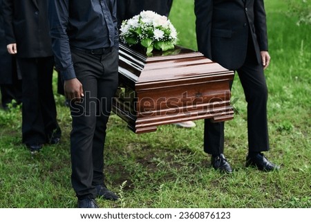 Cropped shot of group of intercultural men in black attire holding by handles of wooden coffin with white flowers on top of closed lid Royalty-Free Stock Photo #2360876123