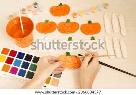 Preparing for Halloween.  On the table there are crafts made from salt dough, pumpkins and monster fingers, multi-colored paints and brushes.  Women's hands paint a pumpkin.  Children's creativity.