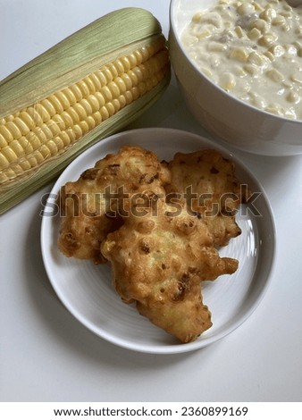 This is a picture of fried corn fritters in a white plate with the uncooked batter and unpeeled corn on a white table.