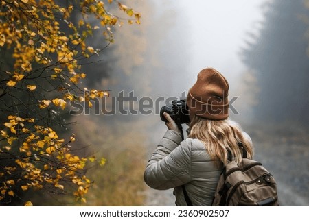 Woman with camera taking picture of autumn leaf. Tourist hiking in misty forest. Landscape photographer with backpack enjoying nature in fall season