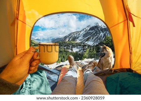 Cup of hot drink in the hand and wonderful view of snowy mountain tops through the open entrance of the tent. The beauty of a romantic hike and camping accompanied by a dog.