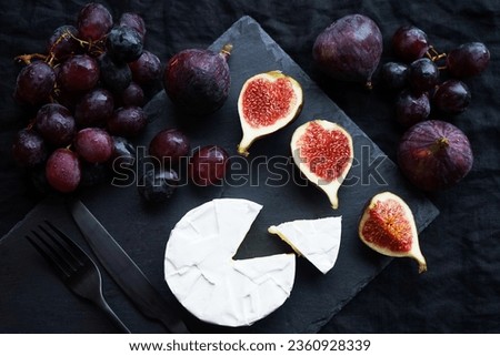 Ripe figs, red grapes with Camembert cheese next to cutlery on a dark background