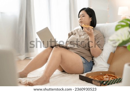 A happy Asian plus-size woman in comfy clothes is using her laptop computer and enjoying her yummy pizza in her bedroom. work from home, freelance, leisure, unhealthy lifestyle