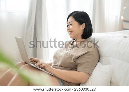 Side view of a happy Asian plus-size woman in comfy clothes is using her laptop computer while relaxing in her bedroom. work from home, freelance, leisure