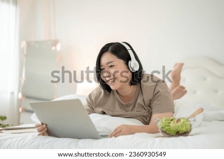 A happy Asian plus-size woman lying on bed, wearing headphones, and using her laptop computer, watching a movie on bed, reading an online blog, talking on video call, or working from home.