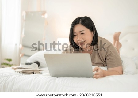 A happy and charming Asian plus-size woman lying on bed and using her laptop computer, watching a movie on bed, reading an online blog, or working from home.