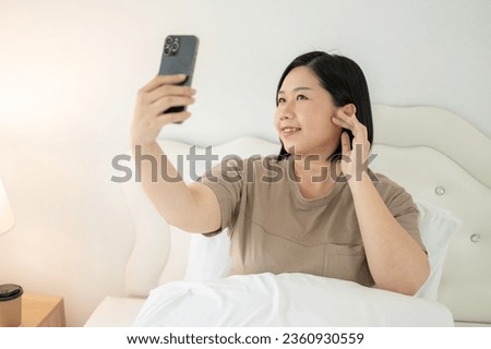 An attractive Asian plus-size woman in comfy clothes is taking selfies on her smartphone while relaxing on her bed in her bedroom. People and lifestyle concepts