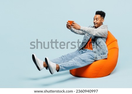 Full body young man of African American ethnicity wear denim jacket orange t-shirt sit in bag chair use play racing app on mobile cell phone gadget for pc video games isolated on plain blue background