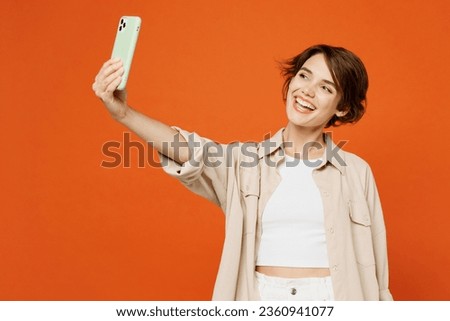 Young happy caucasian woman she wear beige shirt casual clothes doing selfie shot on mobile cell phone post photo on social network isolated on plain orange red background studio . Lifestyle concept