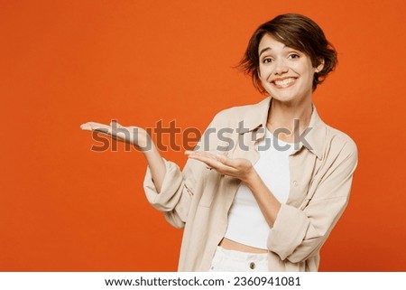 Young happy caucasian woman she wear beige shirt casual clothes point hands arms aside indicate on workspace area copy space mock up isolated on plain orange red background studio. Lifestyle concept