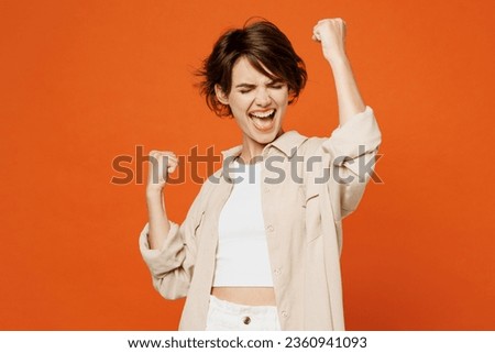 Young excited happy caucasian woman wear beige shirt casual clothes doing winner gesture celebrate clenching fists say yes isolated on plain orange red background studio portrait. Lifestyle concept Royalty-Free Stock Photo #2360941093