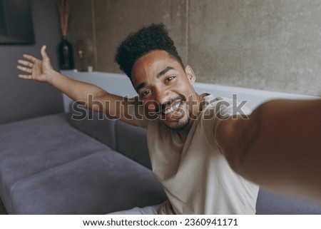 Close up fun young african american man in beige casual t-shirt sit on grey sofa indoors apartment doing selfie shot on mobile phone show flat interior, stretch hand, rest on weekends stay at home.