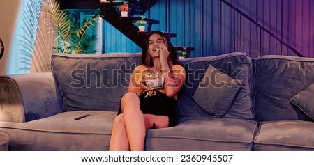 Beautiful young female sitting on sofa watching comedy movie eating popcorn snack enjoy weekend at indoor home. Happy Indian woman laughing looking funny web series on television spend free time