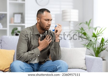 A young African American man is sitting on the couch at home and having an asthma attack. Holds his chest and breathes hard, blows an inhaler into his mouth. Royalty-Free Stock Photo #2360957111