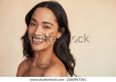 Portrait of young mixed race smiling woman applying foundation swatches of various tone on cheek. Beautiful hispanic woman with different shades of foundation on face isolated against background. Royalty-Free Stock Photo #2360967361