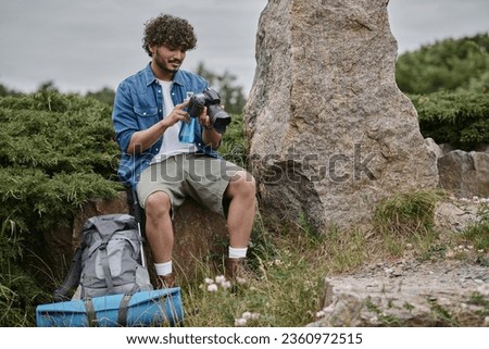 travel and nature concept, indian man holding bottle and using digital camera while sitting on rock