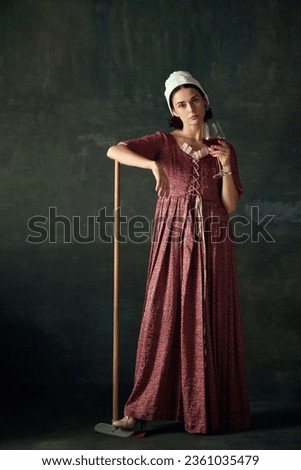 Young emotionless woman in image of renaissance maid standing with mop and drinking red wine against vintage green background. Concept of history, comparison of eras, beauty, art, creativity
